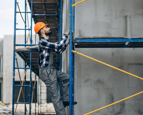 Un lavoratore edile di F.G. Project Sardegna, con elmetto arancione, tuta grigia e camicia a quadri, sale su un ponteggio blu durante un intervento di ristrutturazione o costruzione in un cantiere a Cagliari. Sullo sfondo si vede un edificio residenziale.