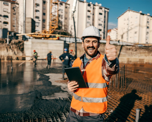 Un tecnico della FG Project Sardegna con elmetto bianco e gilet arancione sorride facendo il segno OK. In mano tiene un tablet per il monitoraggio digitale in un cantiere edile durante la fase di getto delle fondazioni.