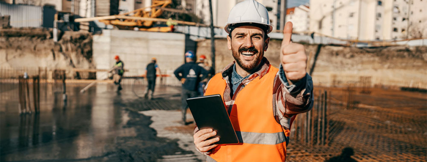 Un tecnico della FG Project Sardegna con elmetto bianco e gilet arancione sorride facendo il segno OK. In mano tiene un tablet per il monitoraggio digitale in un cantiere edile durante la fase di getto delle fondazioni.