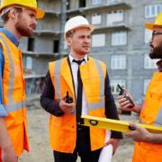 Coordinamento e gestione della sicurezza nei subappalti a cascata in cantiere Tre professionisti del settore edile con caschetti e giubbotti ad alta visibilità si confrontano in cantiere tenendo in mano planimetrie e strumenti di verifica.