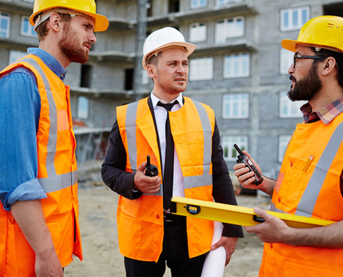 Tre professionisti del settore edile con caschetti e giubbotti ad alta visibilità si confrontano in cantiere tenendo in mano planimetrie e strumenti di verifica.