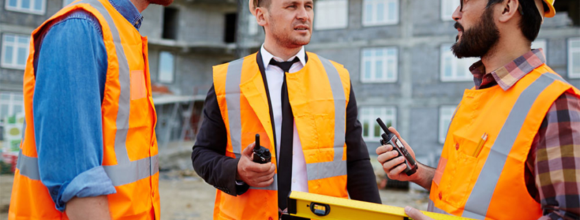Tre professionisti del settore edile con caschetti e giubbotti ad alta visibilità si confrontano in cantiere tenendo in mano planimetrie e strumenti di verifica.