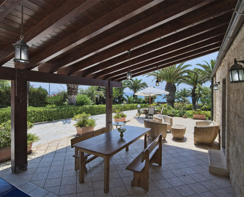 Un ampio pergolato in legno scuro con travi a vista integrato in un agriturismo con pareti in pietra in Sardegna. La struttura offre ombra a un'area relax esterna con tavoli e divani, circondata da piante mediterranee e palme con vista mare.