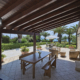 Un ampio pergolato in legno scuro con travi a vista integrato in un agriturismo con pareti in pietra in Sardegna. La struttura offre ombra a un'area relax esterna con tavoli e divani, circondata da piante mediterranee e palme con vista mare.
