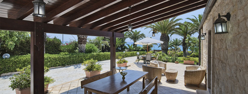 Un ampio pergolato in legno scuro con travi a vista integrato in un agriturismo con pareti in pietra in Sardegna. La struttura offre ombra a un'area relax esterna con tavoli e divani, circondata da piante mediterranee e palme con vista mare.