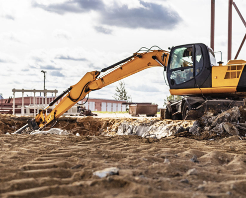 Un escavatore cingolato giallo operativo in un cantiere edile su terreno sabbioso, con il braccio meccanico esteso per operazioni di scavo. L'immagine evidenzia l'importanza della visibilità e delle tecnologie di sicurezza per le manovre di retromarcia.