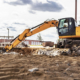 Un escavatore cingolato giallo operativo in un cantiere edile su terreno sabbioso, con il braccio meccanico esteso per operazioni di scavo. L'immagine evidenzia l'importanza della visibilità e delle tecnologie di sicurezza per le manovre di retromarcia.