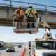 Due operatori con dispositivi di protezione individuale (DPI) al lavoro nel cestello di una piattaforma aerea (PLE) in autostrada, protetti da segnaletica e coni stradali.