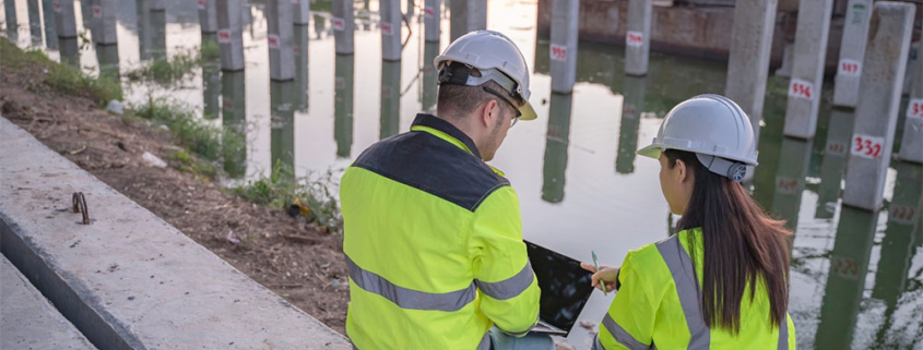 Due tecnici esperti in sicurezza sul lavoro con casco e giubbotti ad alta visibilità analizzano i dati al computer portatile, seduti all'interno di un cantiere edile vicino a un'area parzialmente allagata con pilastri in cemento.