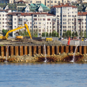 Fotografia aerea di un cantiere edile costiero con palancole metalliche che sputano acqua. Un escavatore giallo opera su terreno instabile vicino al mare in una zona urbana affollata. Illustrazione del rischio idrogeologico e della sicurezza nei cantieri complessi gestita da FG Project Sardegna.