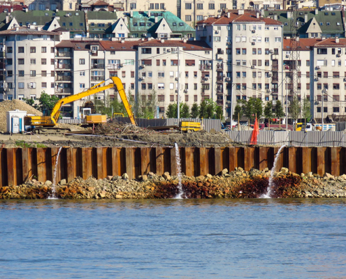 Fotografia aerea di un cantiere edile costiero con palancole metalliche che sputano acqua. Un escavatore giallo opera su terreno instabile vicino al mare in una zona urbana affollata. Illustrazione del rischio idrogeologico e della sicurezza nei cantieri complessi gestita da FG Project Sardegna.