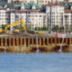 Fotografia aerea di un cantiere edile costiero con palancole metalliche che sputano acqua. Un escavatore giallo opera su terreno instabile vicino al mare in una zona urbana affollata. Illustrazione del rischio idrogeologico e della sicurezza nei cantieri complessi gestita da FG Project Sardegna.