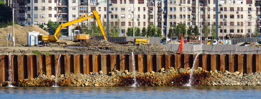 Fotografia aerea di un cantiere edile costiero con palancole metalliche che sputano acqua. Un escavatore giallo opera su terreno instabile vicino al mare in una zona urbana affollata. Illustrazione del rischio idrogeologico e della sicurezza nei cantieri complessi gestita da FG Project Sardegna.